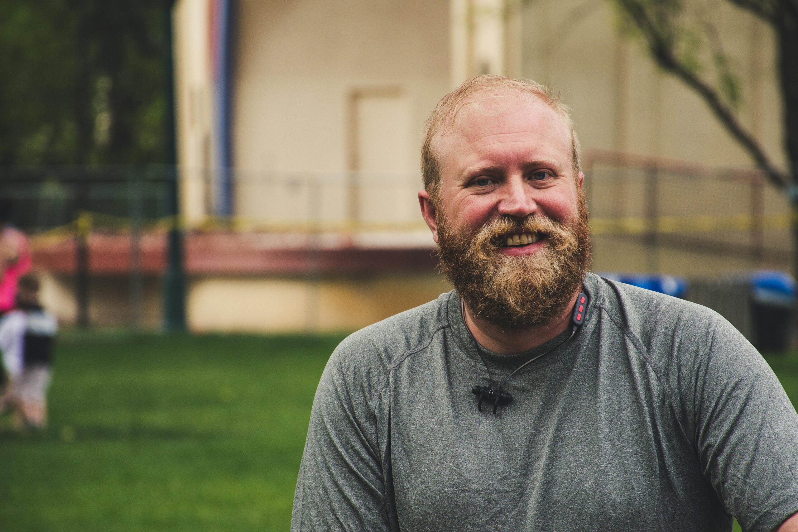 A cheerful bearded man smiling outdoors in a casual setting with greenery.